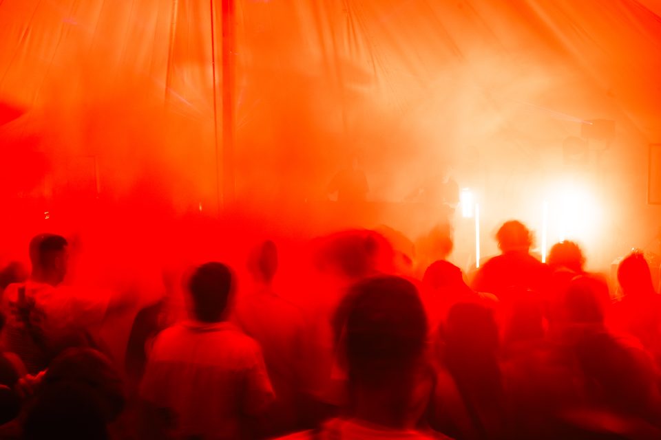 Audience silhouettes under red stage lighting, with performers visible in haze.