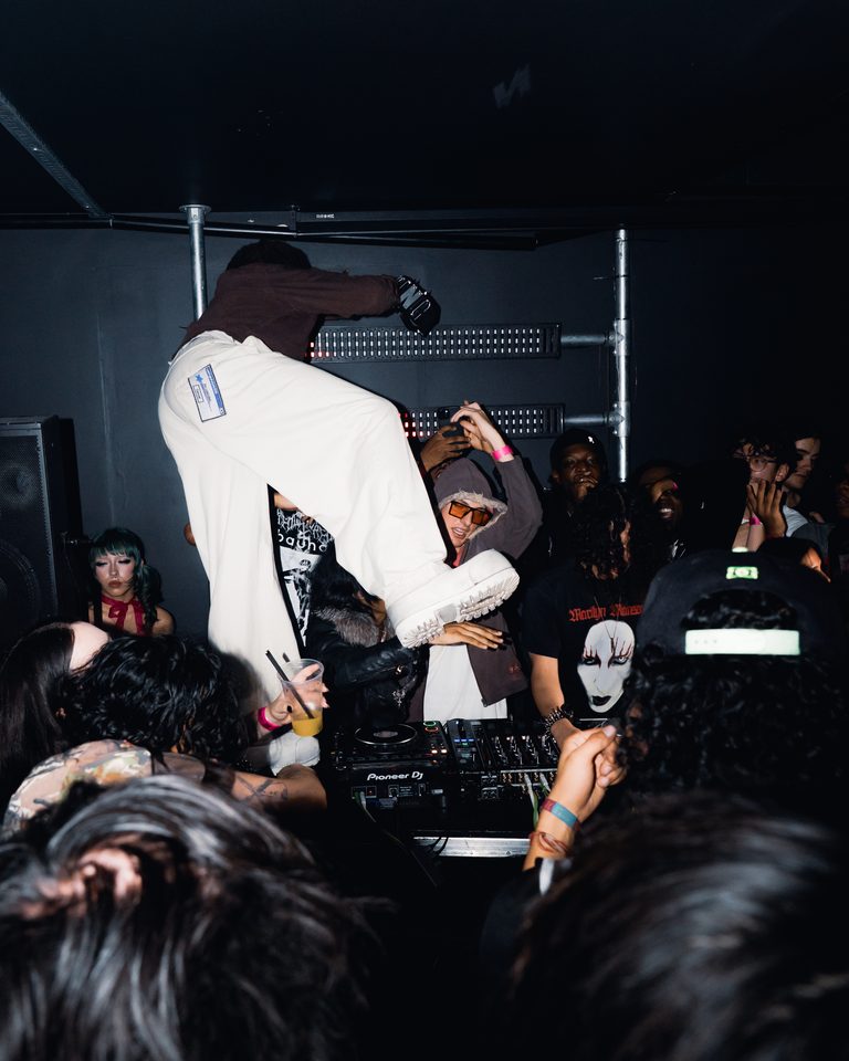 A DJ in a booth with another performer nearby, surrounded by a crowd in a dark club.
