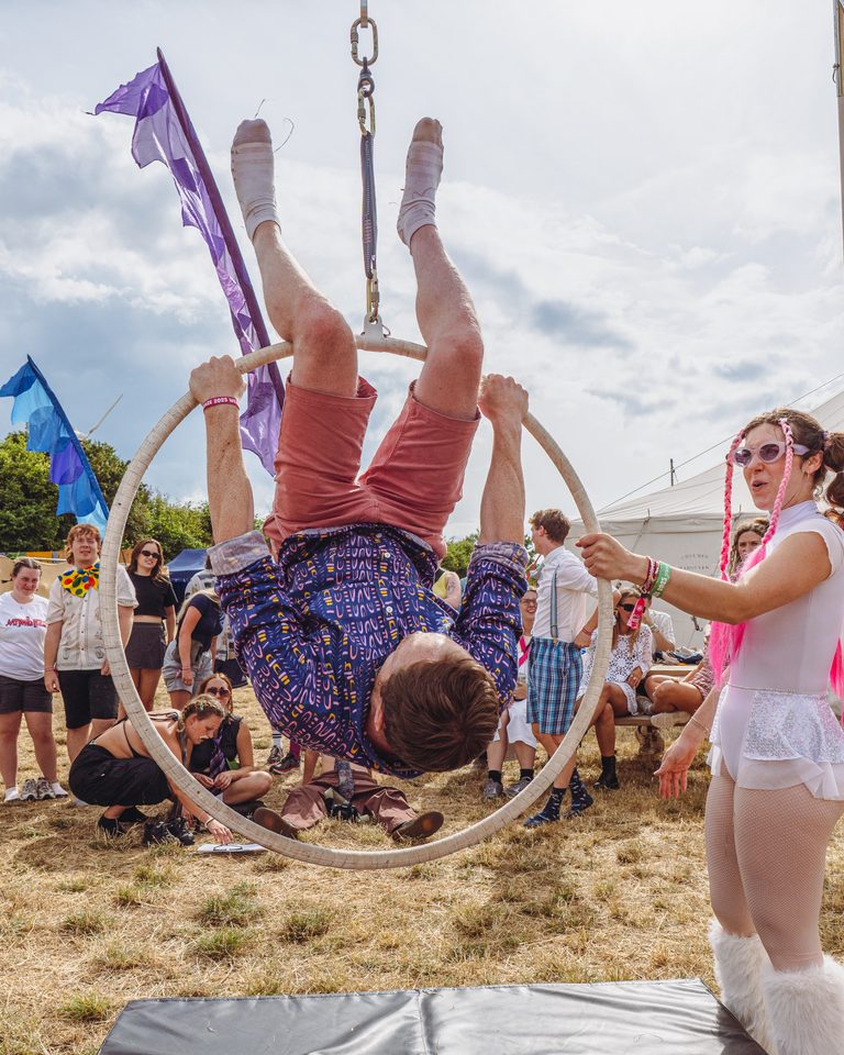 Aerialist hangs upside down in a hoop, surrounded by spectators under daylight.