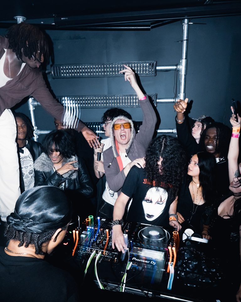 A DJ with equipment surrounded by a crowd in a dimly lit club.