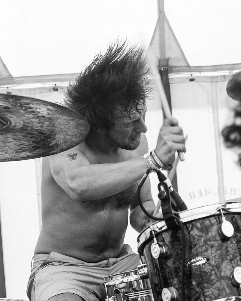 A shirtless drummer with wild hair plays drums under natural light.