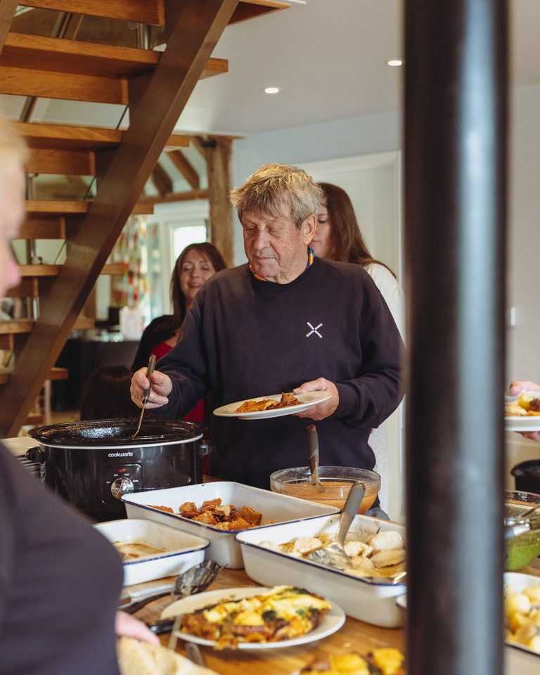 Person serving food from slow cooker at indoor buffet with guests in background.