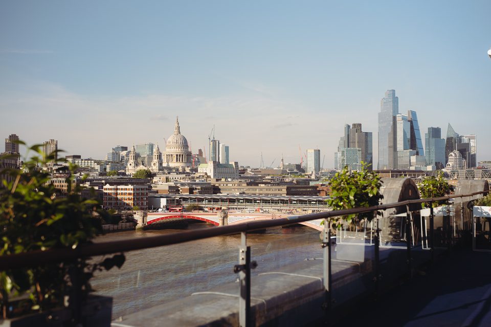 Sunlit urban skyline with river and bridge, framed by railing and foliage.