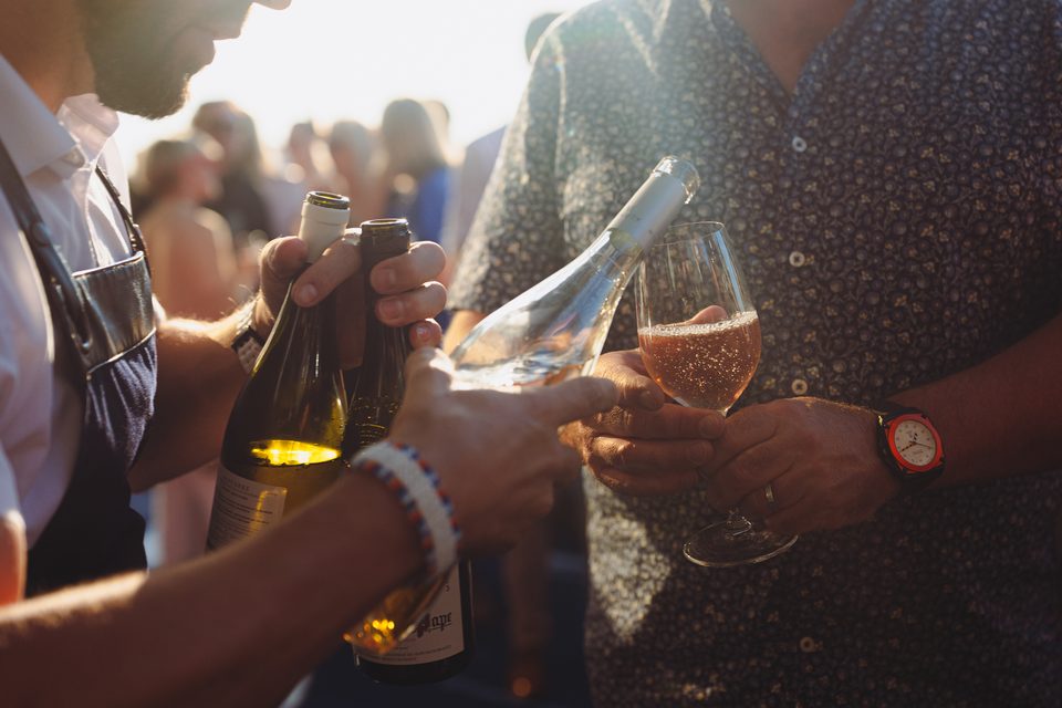 Two people pouring wine into a glass, with a crowd blurred in background.