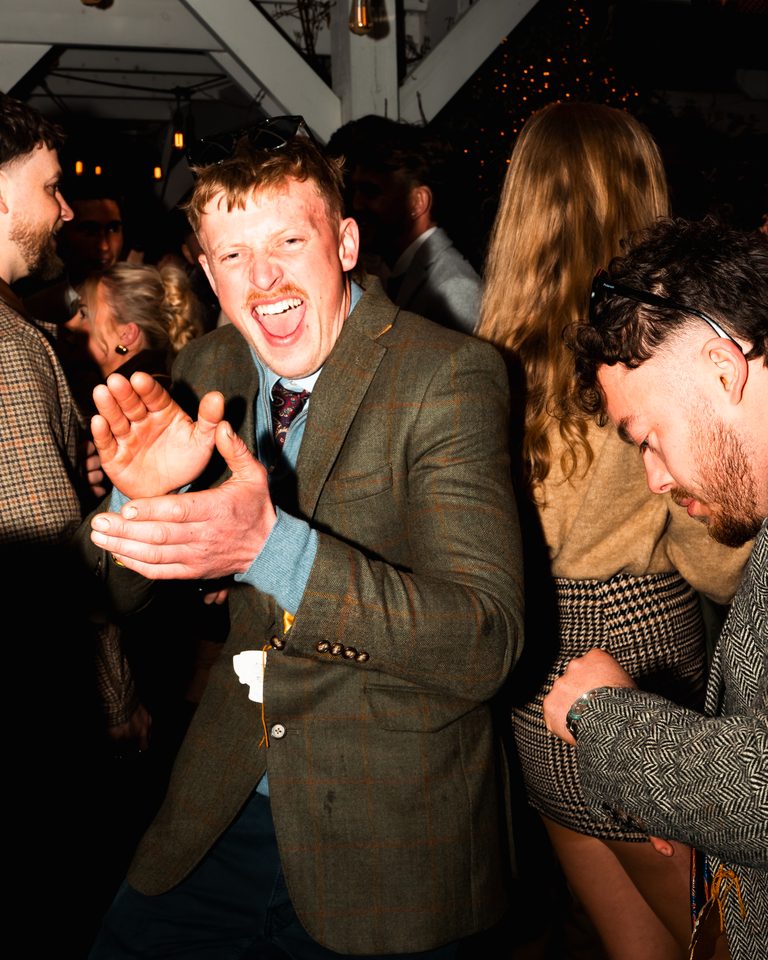 Person in tweed jacket laughing with hands open, surrounded by others in dimly lit indoor space.