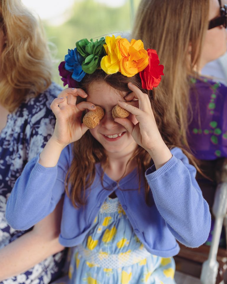 Girl wearing rainbow flower crown holds corks to eyes, smiling.