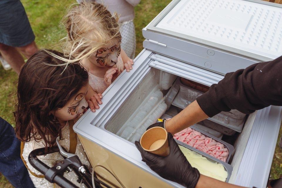 Two children with face paint peer into an open freezer as a vendor serves ice cream.