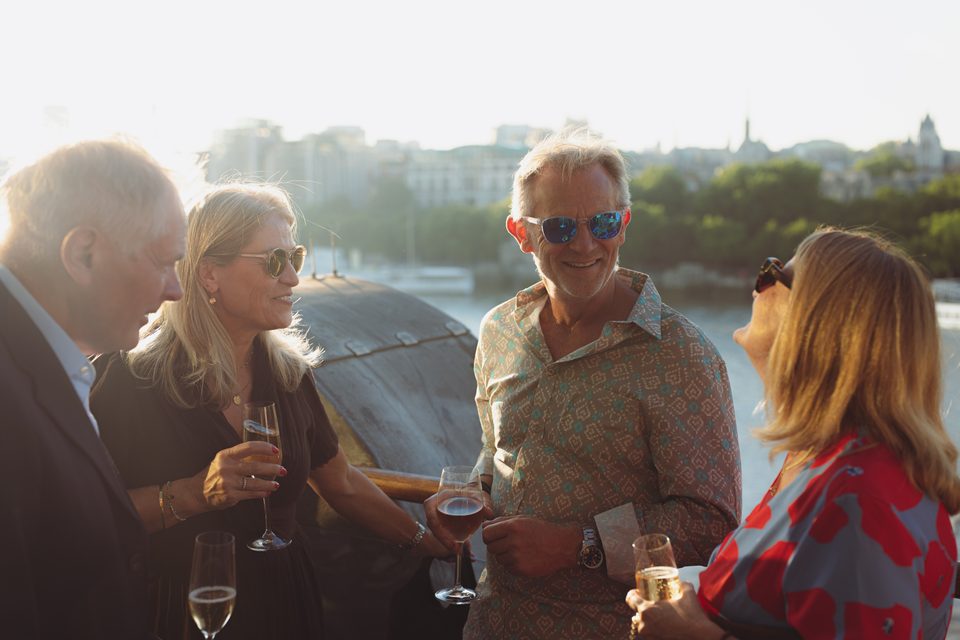 Four people socializing with drinks, sunlit, near water, cityscape background.