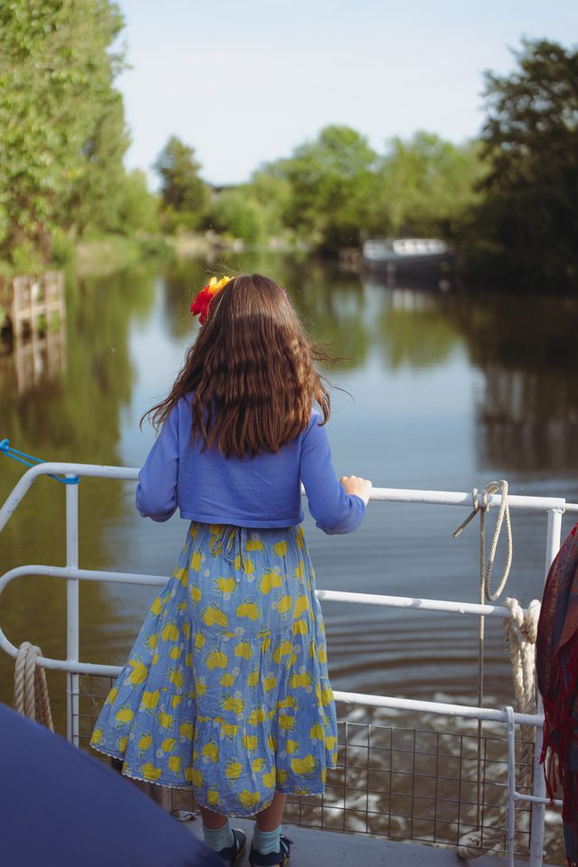 A young girl in a floral dress stands on a boat railing, gazing at a calm river surrounded by greenery.