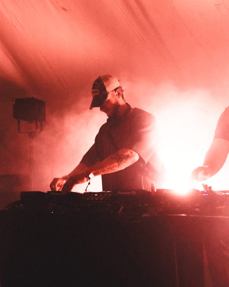 A DJ in a red-lit booth with smoke, hands on equipment, backlit.