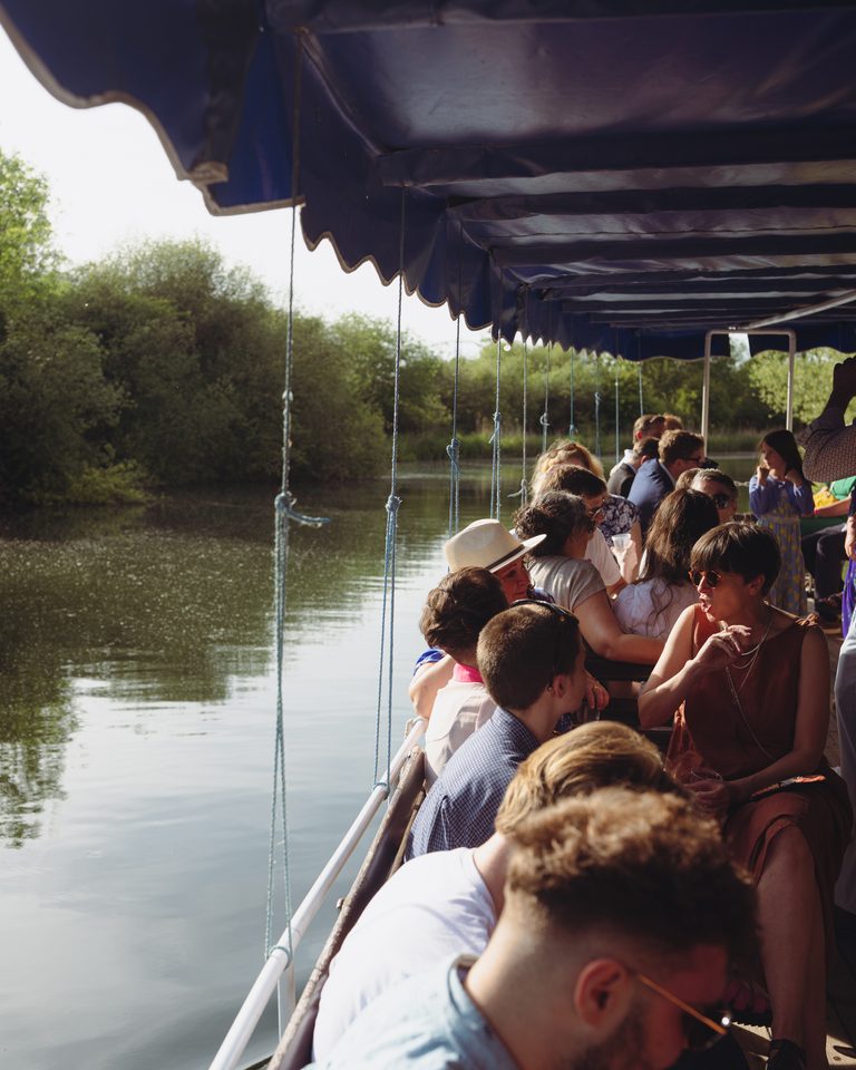 Passengers seated under blue canopy on riverboat, surrounded by greenery and water.