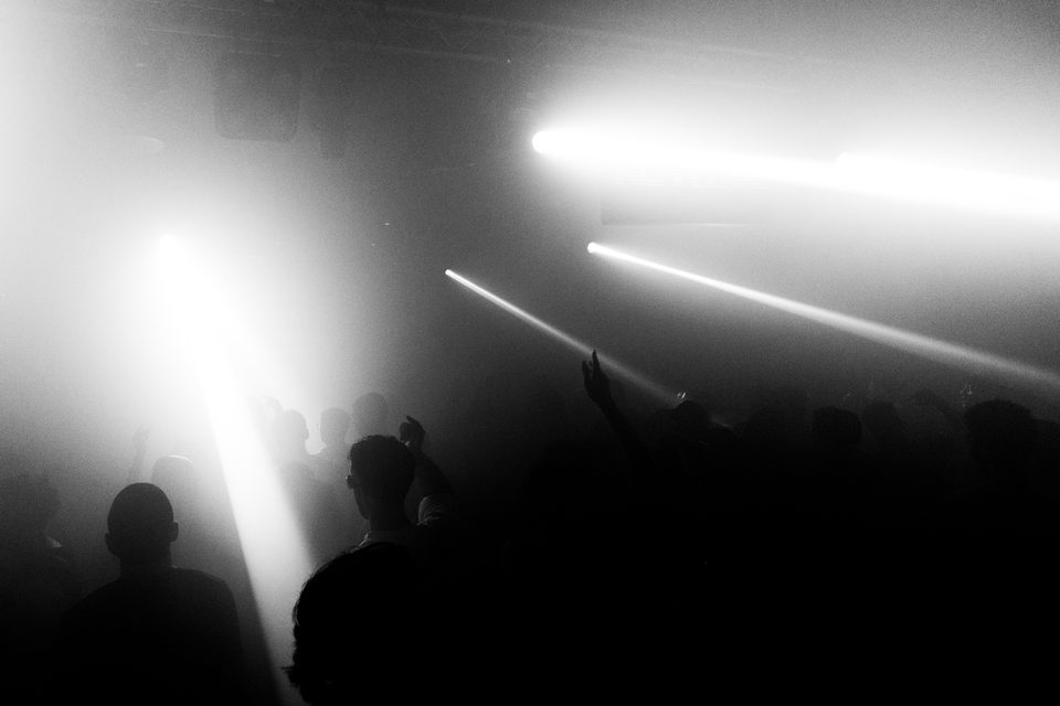 Silhouetted crowd in a dim club with bright stage beams cutting through haze.