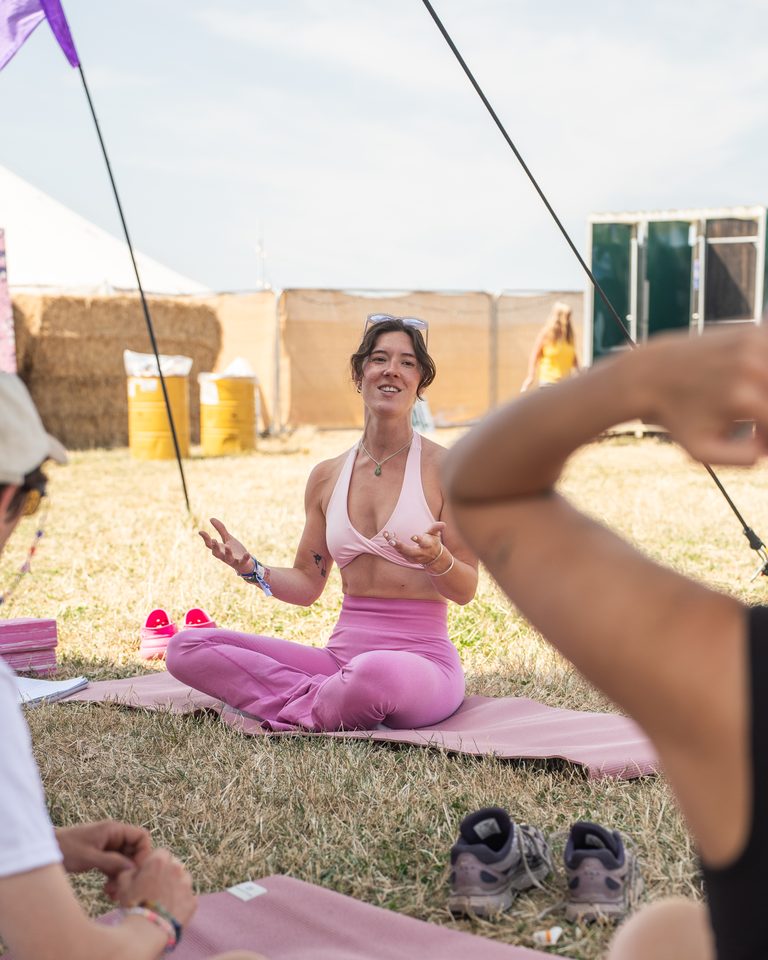 Person in pink yoga attire sits on mat outdoors, smiling, with others partially visible.