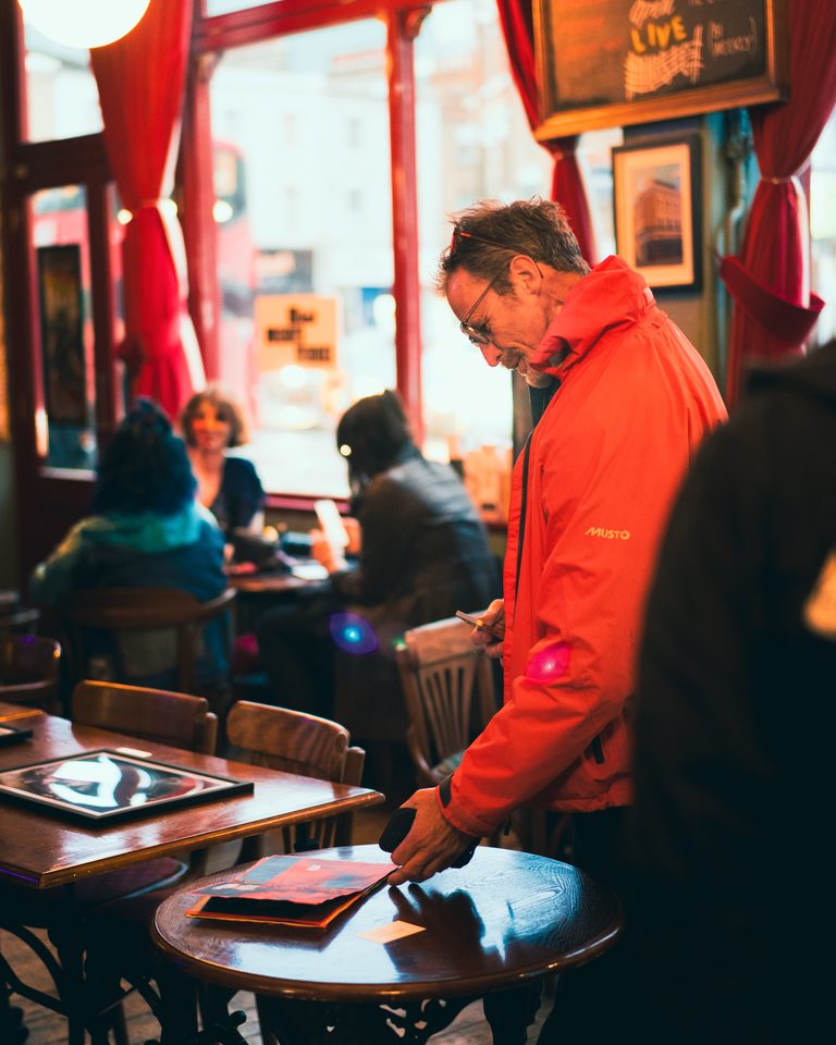 Person in red jacket at small table, others seated nearby in dimly lit interior.