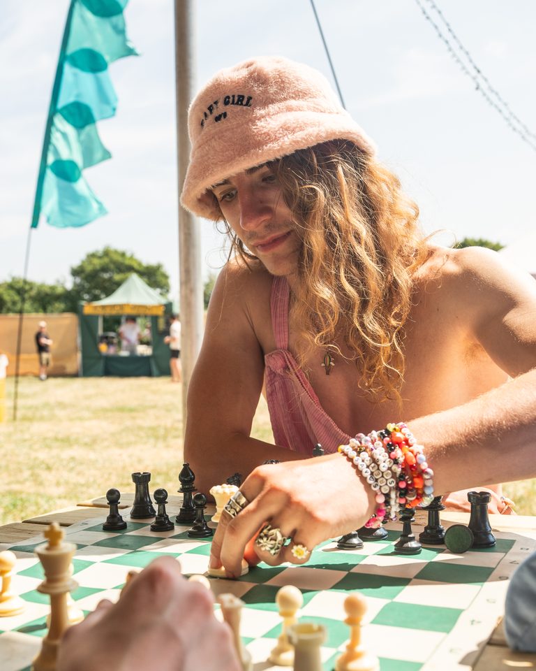 Person playing chess outdoors under bright sun, wearing pink hat and colorful bracelets.