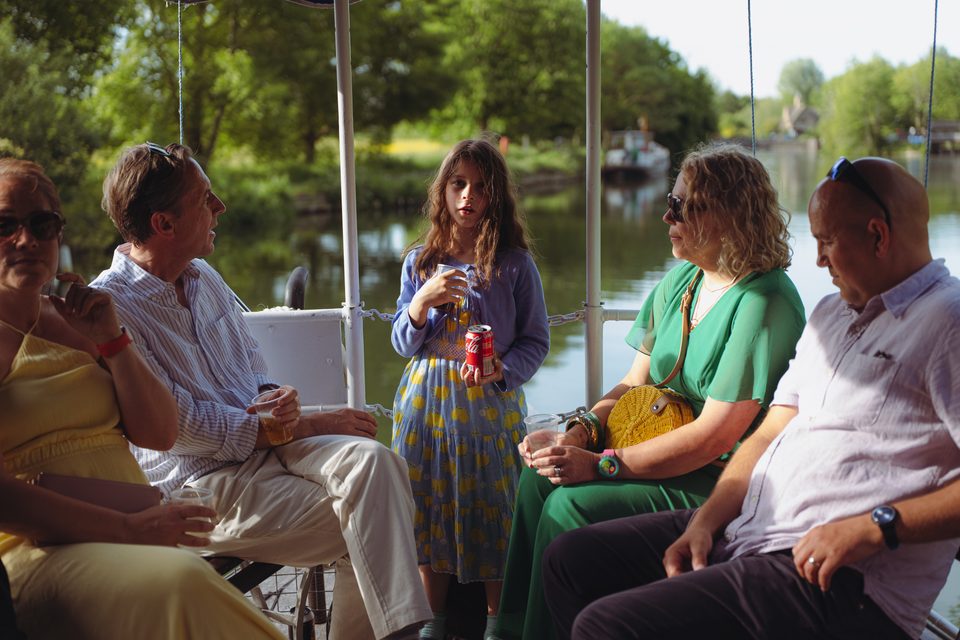 Five people on a boat, some seated, one standing, holding drinks, under natural light.