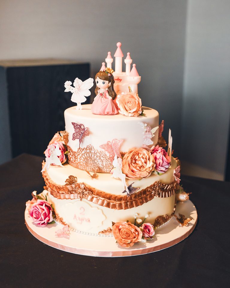 A three-tiered birthday cake with pink roses, butterflies, and a castle topper.