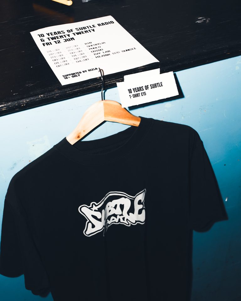 Black T-shirt with white logo on wooden hanger, price tag visible, event flyer above.