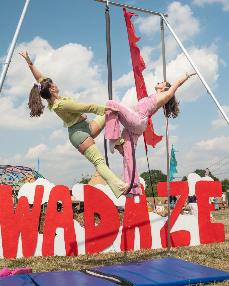 Two performers suspended in mid-air on aerial apparatus against a bright sky.