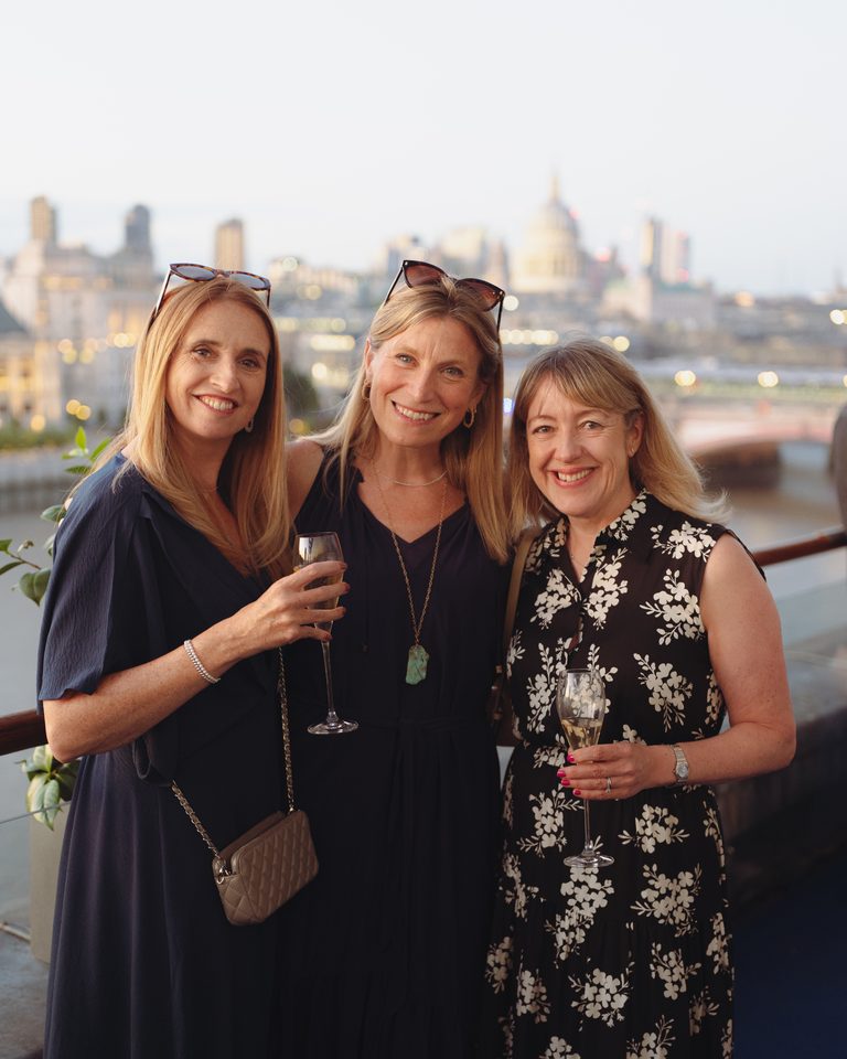 Three women in evening wear holding champagne glasses, smiling, with city skyline in soft focus behind them.