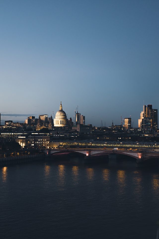 Evening view of city skyline with illuminated buildings and river reflections.