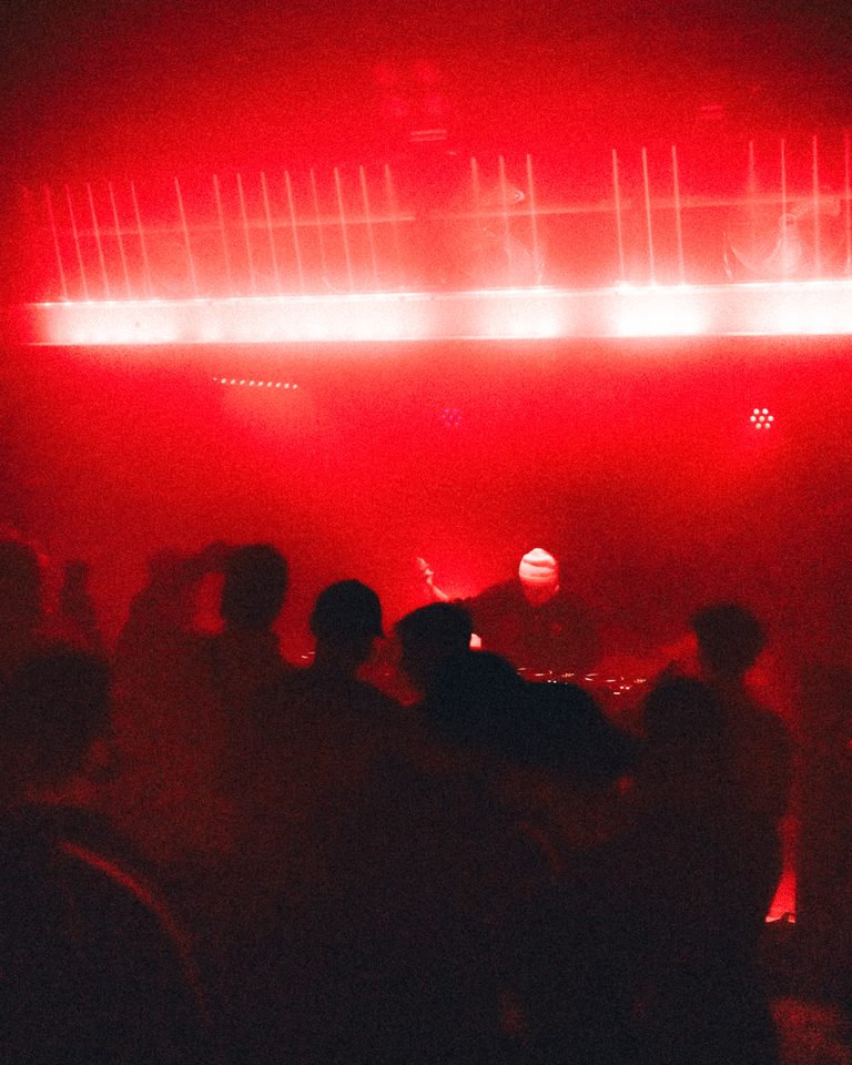 Silhouetted crowd facing a DJ under red stage lights.