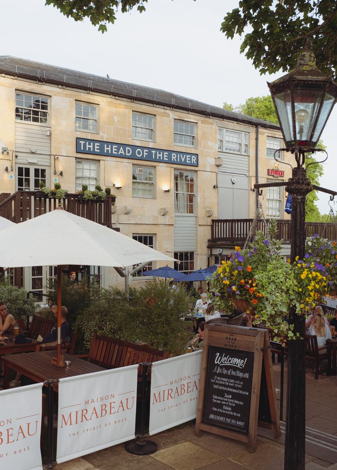 People seated at tables under umbrellas, with a pub sign and hanging flowers.