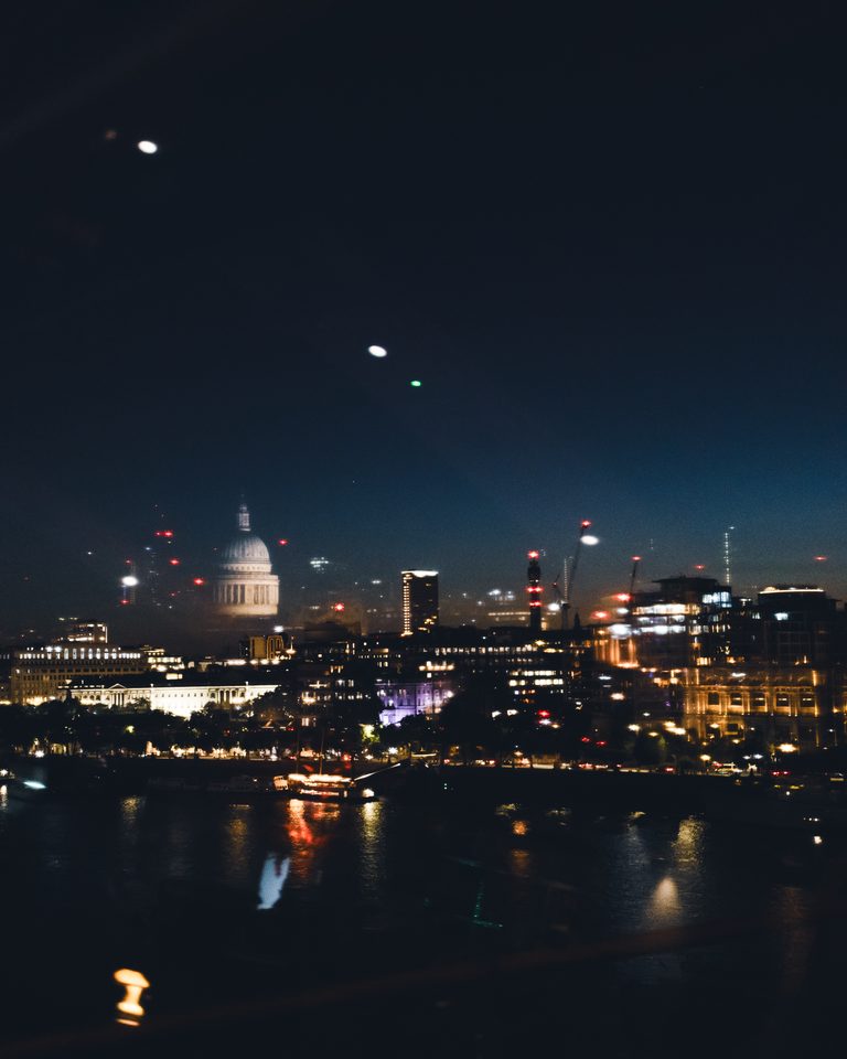 Night view of a city skyline with illuminated buildings and a river, featuring a prominent dome and scattered lights.