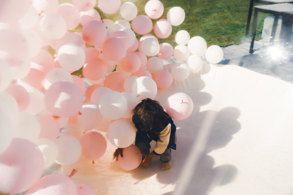 Child playing with pink and white balloons under bright sunlight.