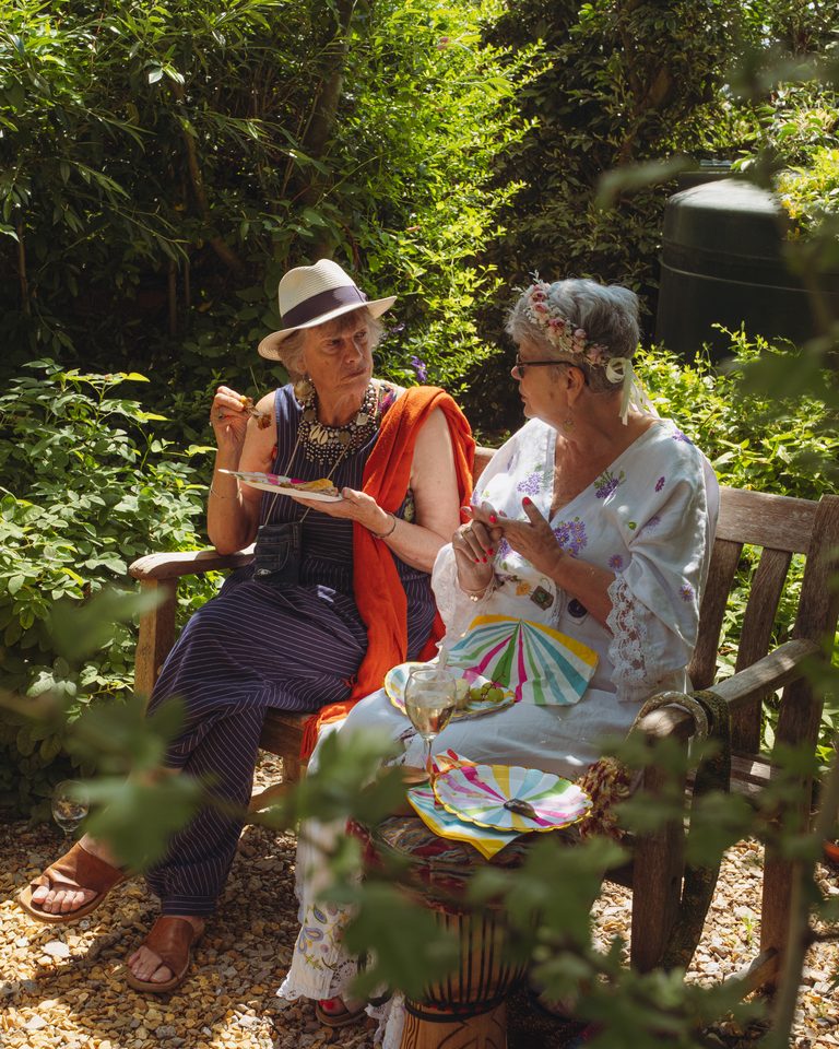 Two women seated on a bench in a sunlit garden, eating and conversing.