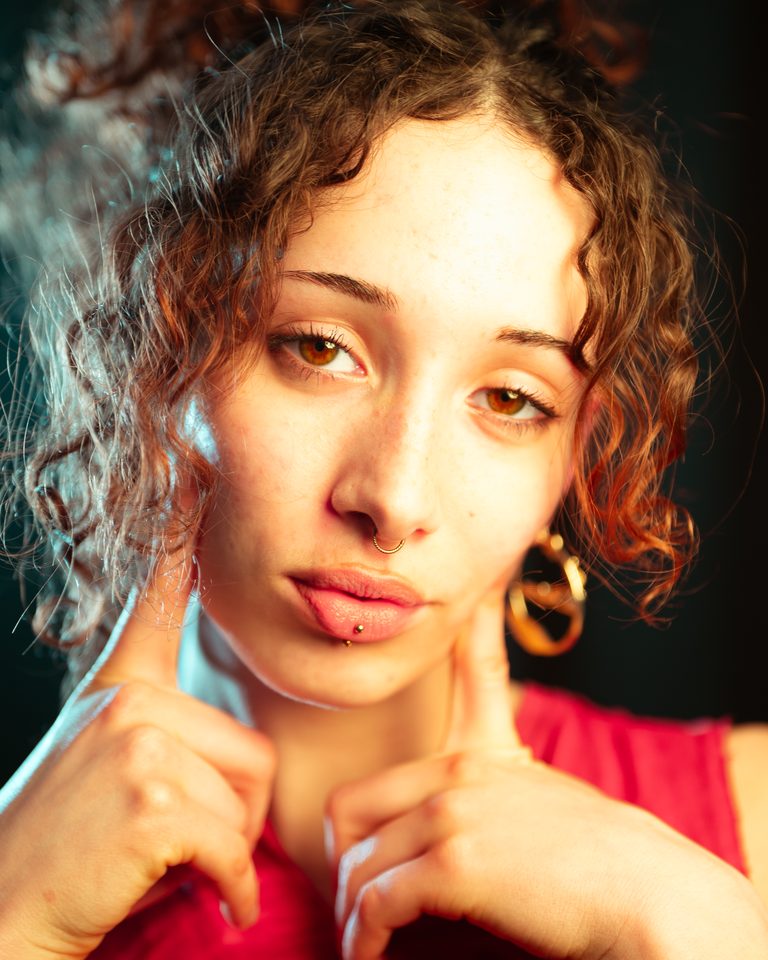 Portrait of a person with curly hair and piercings, wearing a red top.