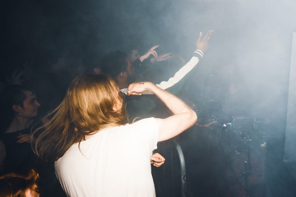 A performer with long hair and a white shirt raises a hand in a smoky club environment.