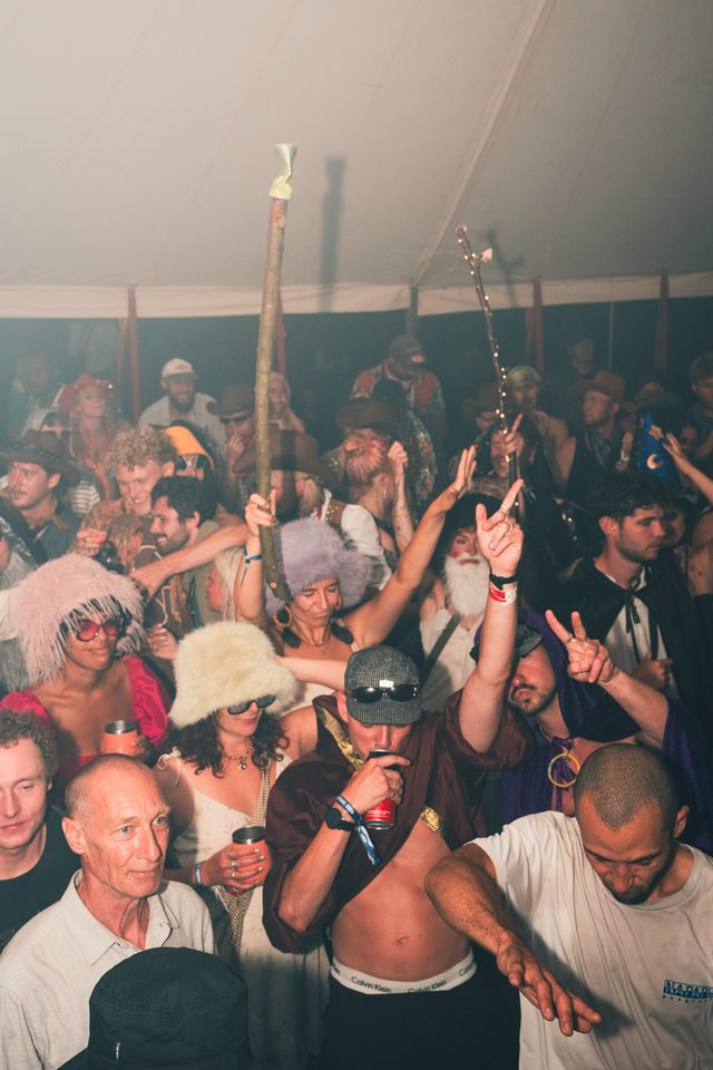 Diverse group of people in costumes under dim lighting, some holding sticks or making hand gestures.