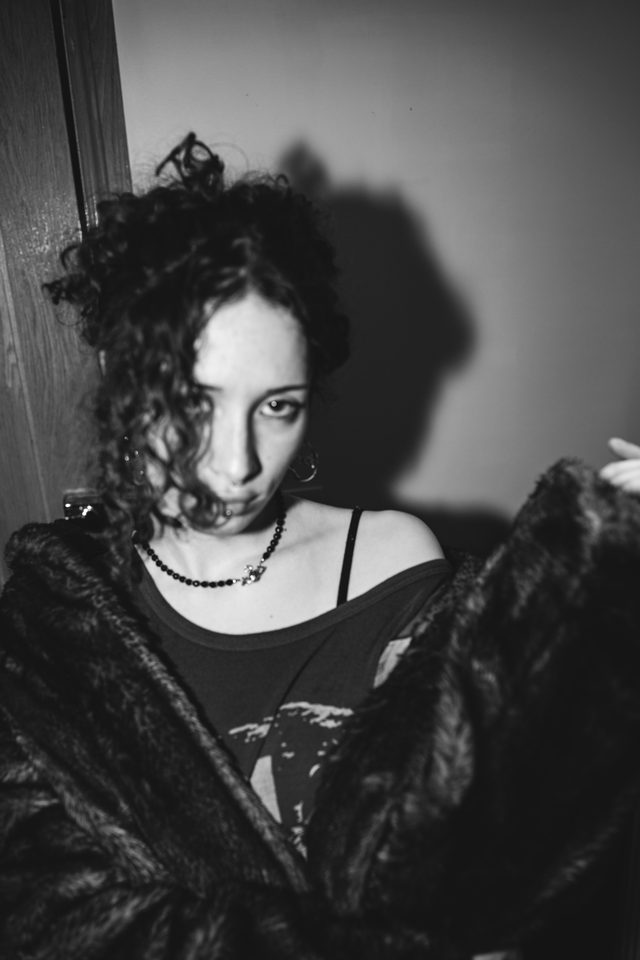 A person in a fur coat, wearing a beaded necklace, against a plain wall.