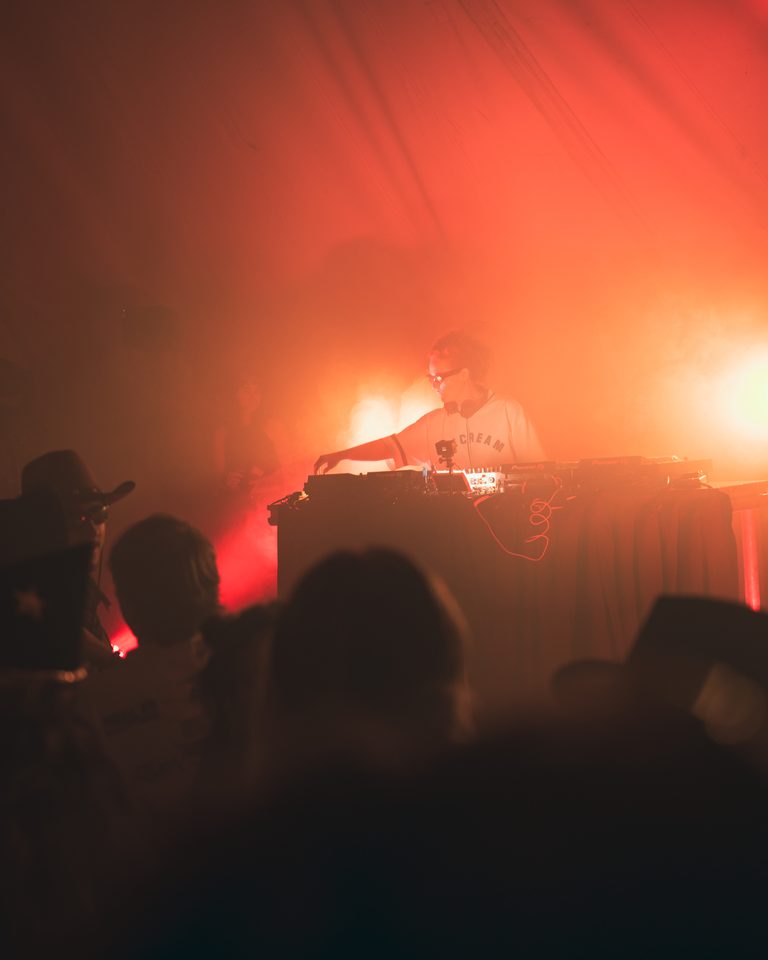 DJ behind console under red stage light, crowd in silhouette, smoke haze.