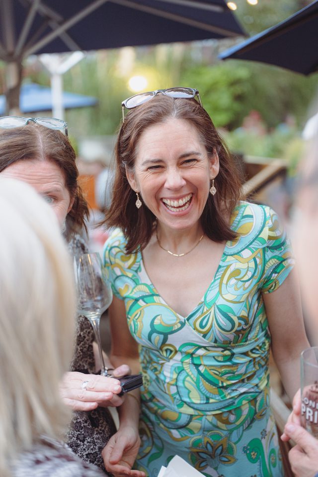 Two women at an outdoor event under umbrellas, one laughing with glasses on head.