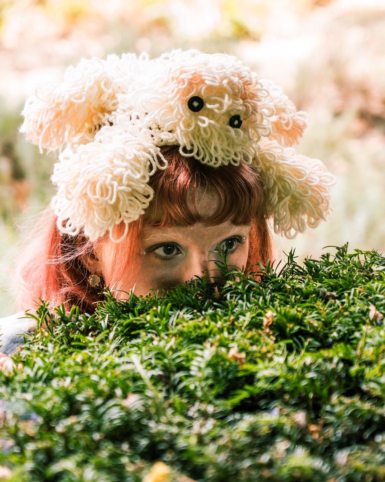 A person with a knitted hat peers from behind a bush.