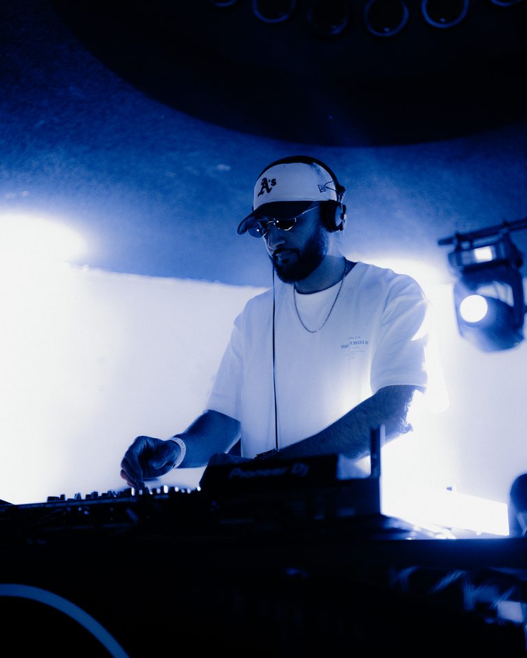 A DJ wearing a cap and headphones, illuminated by backlit stage lights.