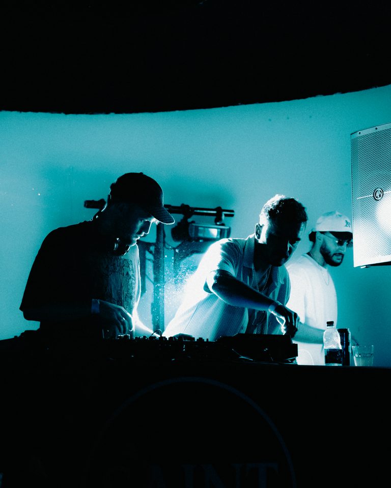 Three performers at DJ booth under blue stage light, silhouetted against bright background.