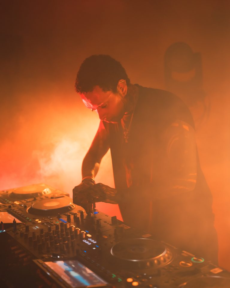 A DJ with tattoos mixes tracks under orange stage lights in a smoky indoor club.