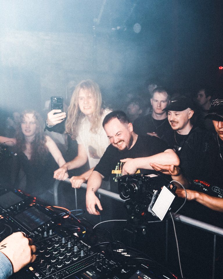 DJ behind equipment with crowd reaching forward in dimly lit club.