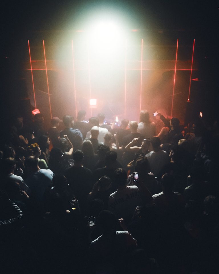 Dense crowd in a dim club, silhouetted against bright stage lights and red laser beams.