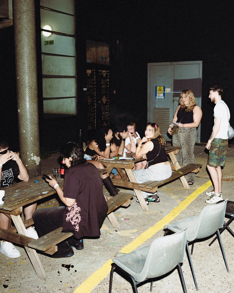 Group of people socializing at outdoor picnic tables under artificial lighting.