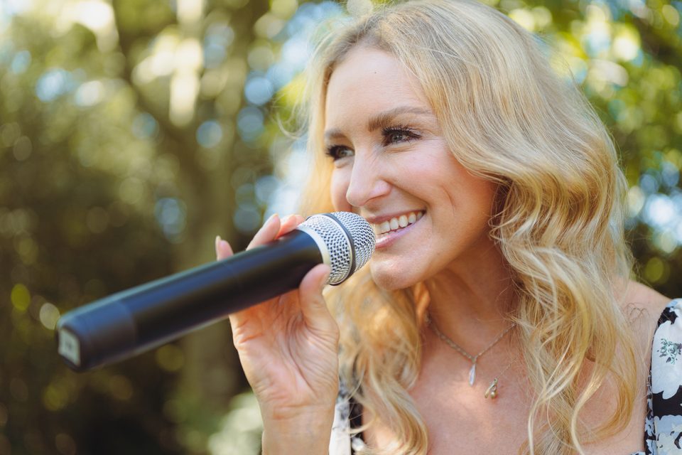 A person with blonde hair holding a microphone, smiling outdoors in daylight.
