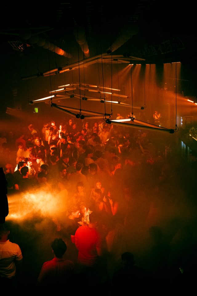 Crowd dancing in a smoky club under red stage lights.