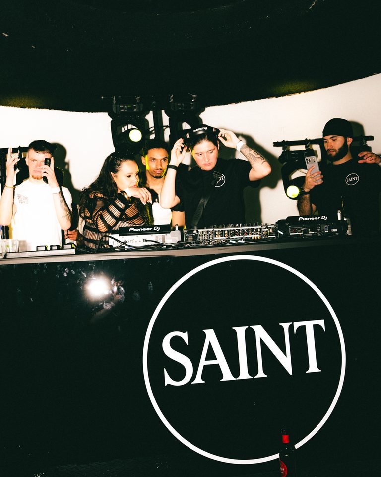 Group of performers and guests at a DJ booth under stage lights.