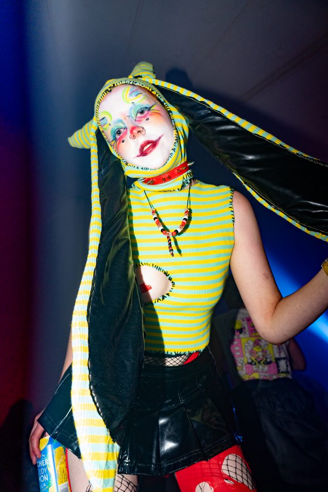 Person in colorful clown makeup and striped outfit, holding drink, under club lighting.