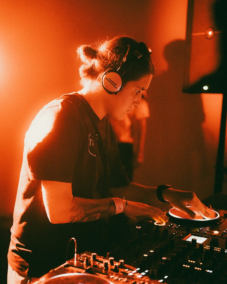 DJ wearing headphones, adjusting controls under red stage lighting.
