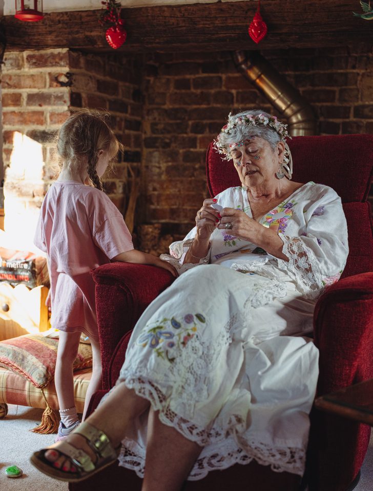 An elderly person in a white dress sits in a red chair, a young girl stands beside her.