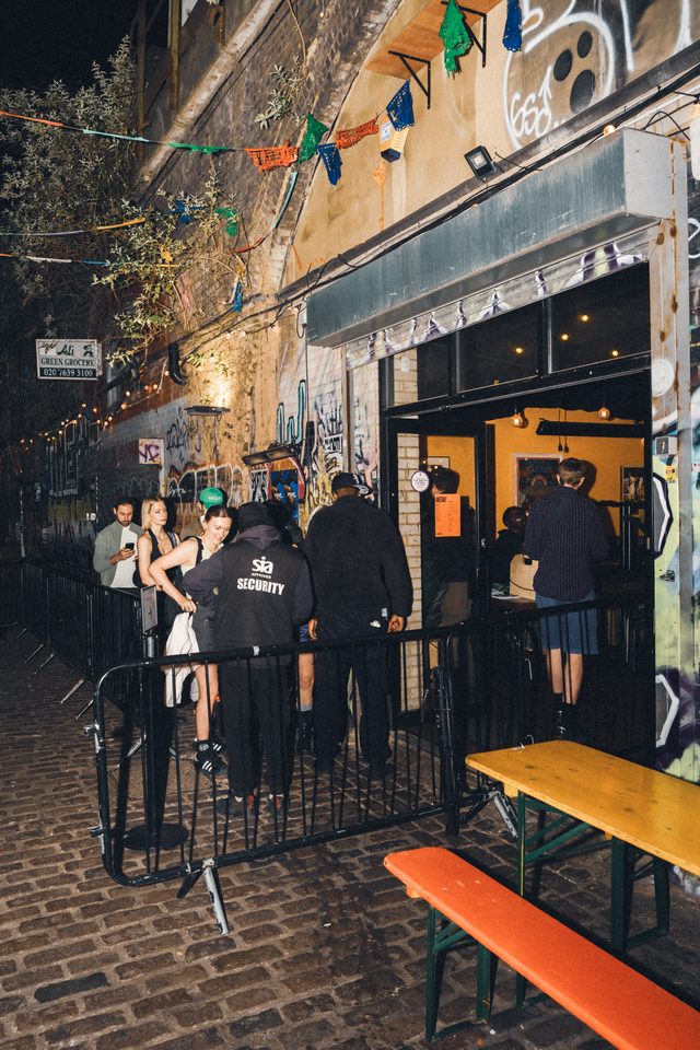 People queueing outside a venue with security, under string lights on cobblestone street.